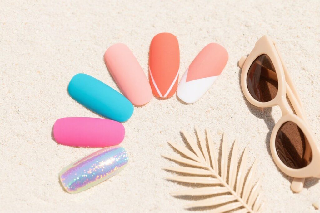 5 different eleglara's press-on nails are laying on the sands. On the side, there is a leaves and a pair of glasses.